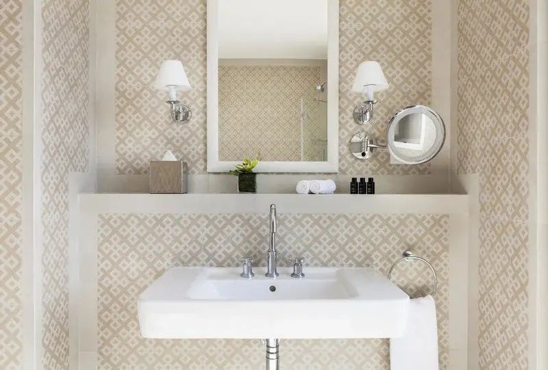 Weißes Waschbecken vor gemusterter Fliesenwand mit großem Spiegel und Wandleuchten.