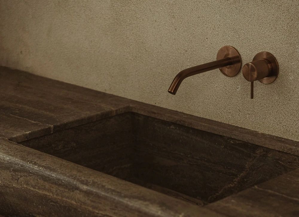 Nahaufnahme von einem dunklen Natursteinwaschbecken mit einer schlichten Wandarmatur aus Bronze