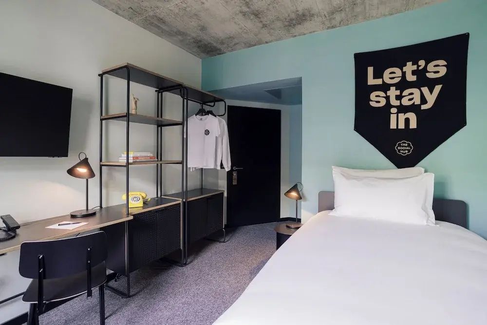 Modernes Hotelzimmer mit weißem Bett, schwarzem Regalsystem, Schreibtisch und hellblauer Wand.