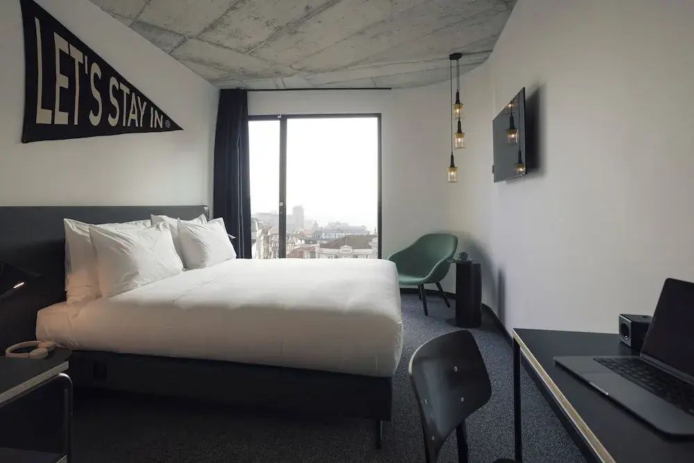 Moderner Schlafbereich mit Doppelbett, grünem Sessel, Fernseher und Blick durch ein Fenster auf die Stadt.