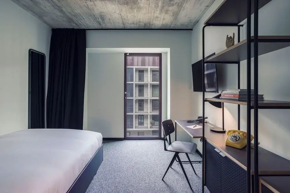 Blick in ein Hotelzimmer mit Bett, Schreibtisch, Stuhl und großem Fenster zum Innenhof.