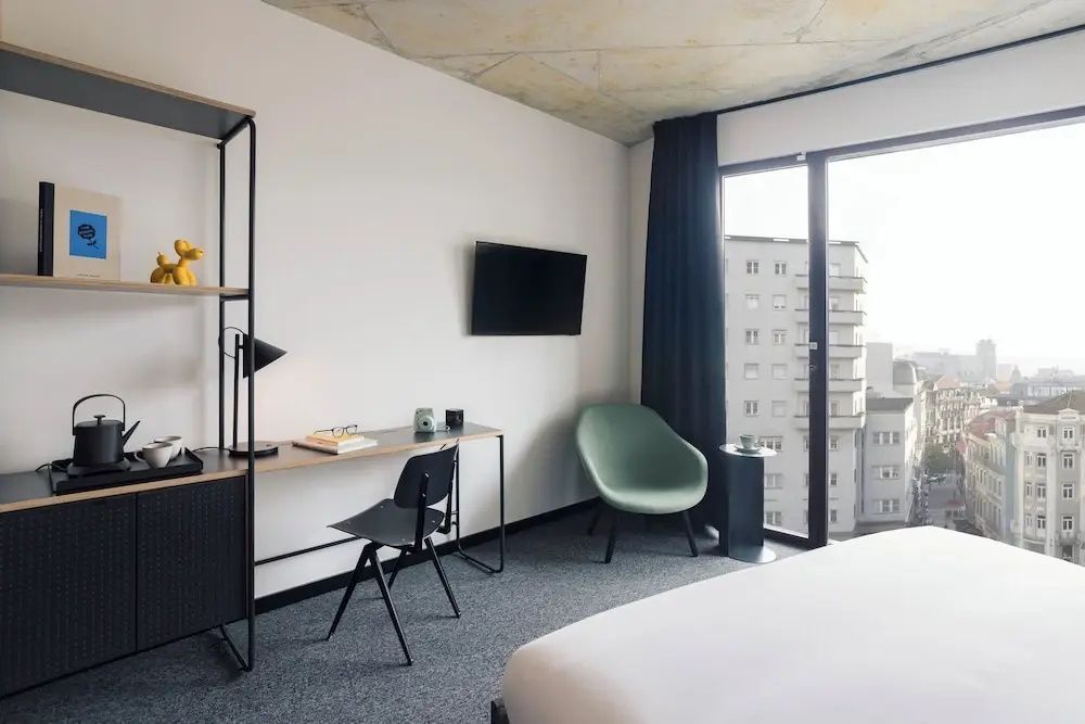 Arbeitsplatz mit Schreibtisch, Stuhl, Regal und grünem Sessel neben einer großen Fensterfront.