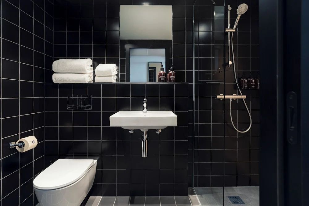 Badezimmer mit schwarzen Fliesen, weißem Waschbecken, Toilette und Dusche mit Glaswand.