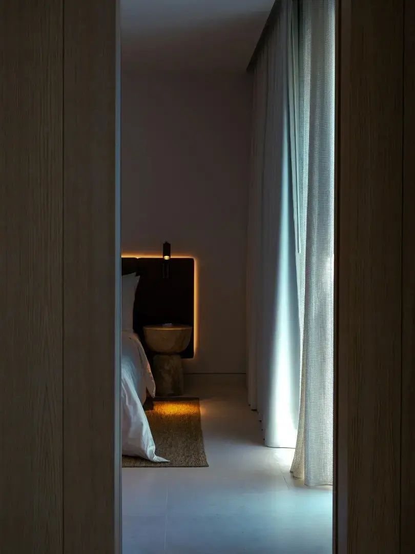 Blick durch den Türspalt auf ein schlichtes Bett mit beleuchtetem Kopfteil und Beistelltisch.