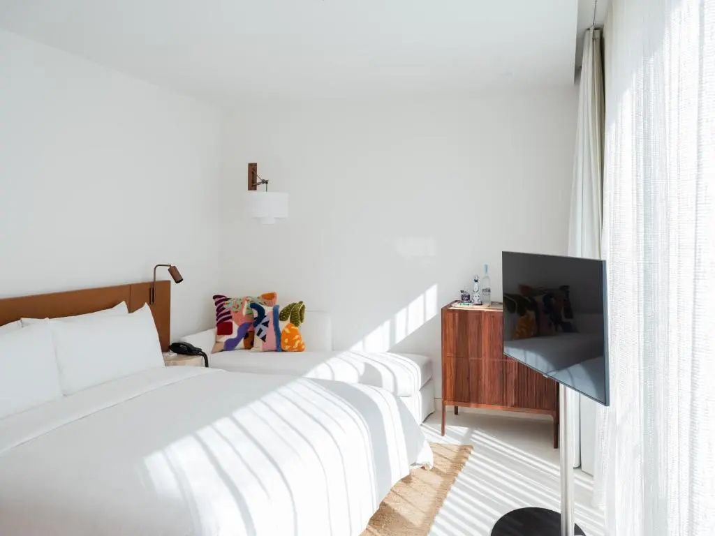 Lichtdurchflutetes Hotelzimmer mit großem Bett, Fernseher auf Ständer und Sitzgelegenheit.