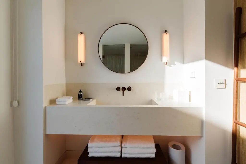 Badezimmer mit rundem Spiegel, wandmontierten Leuchten und hellem Waschtisch mit Handtüchern.