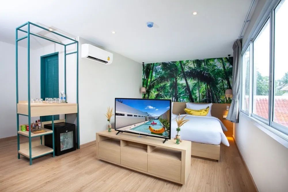 Helles Hotelzimmer mit Doppelbett, Fernseher auf einem Holzschrank und Tropentapete an der Wand.