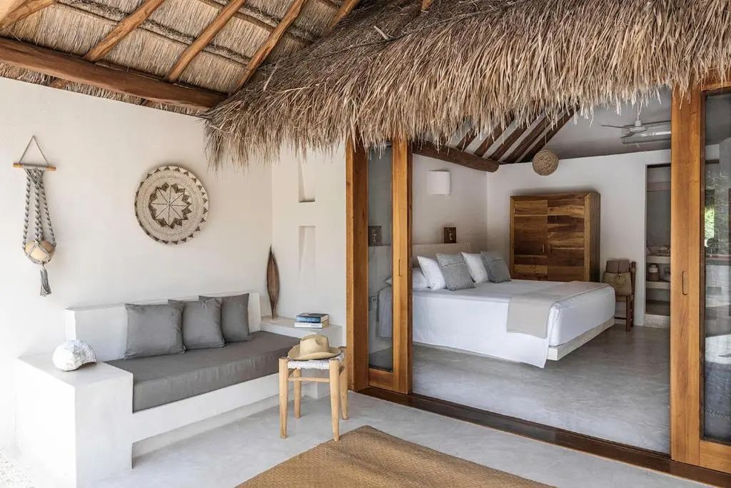 Reetgedeckte Terrasse mit grauem Sofa und offener Schiebetür zum Schlafbereich mit großem Bett.