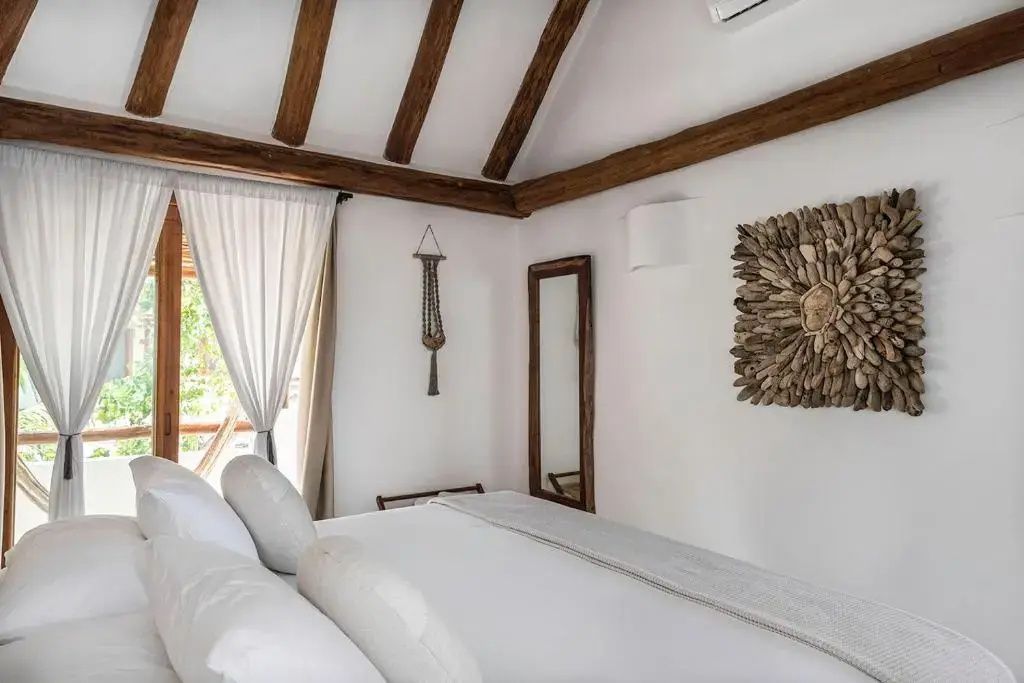 Hell eingerichtetes Hotelzimmer mit Doppelbett, Holzbalken an der Decke und Wandspiegel.
