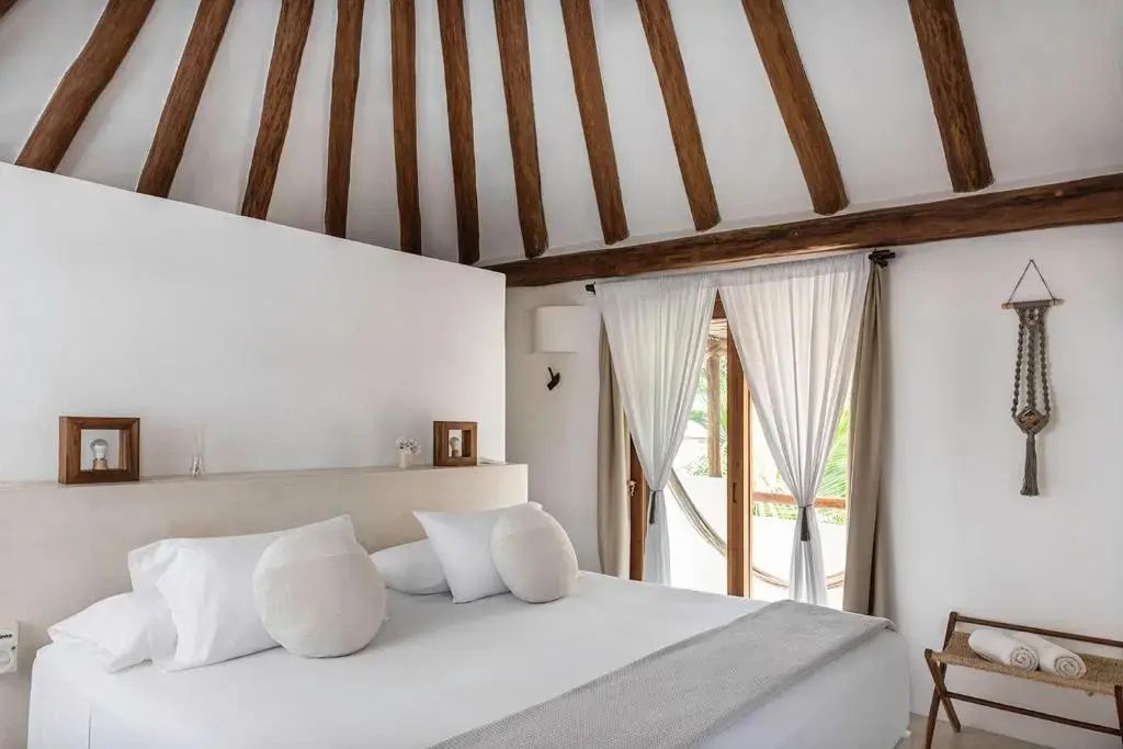 Doppelbett in einem Zimmer mit hoher Holzkonstruktion an der Decke und hellem Vorhang.