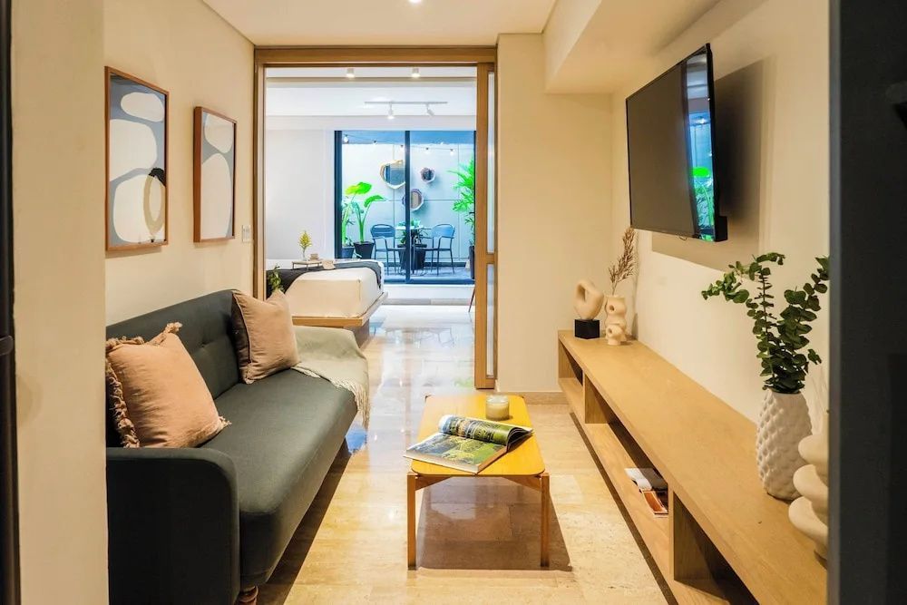 Wohnbereich mit dunkelgrünem Sofa, Holztisch, Fernseher und Blick in ein Schlafzimmer