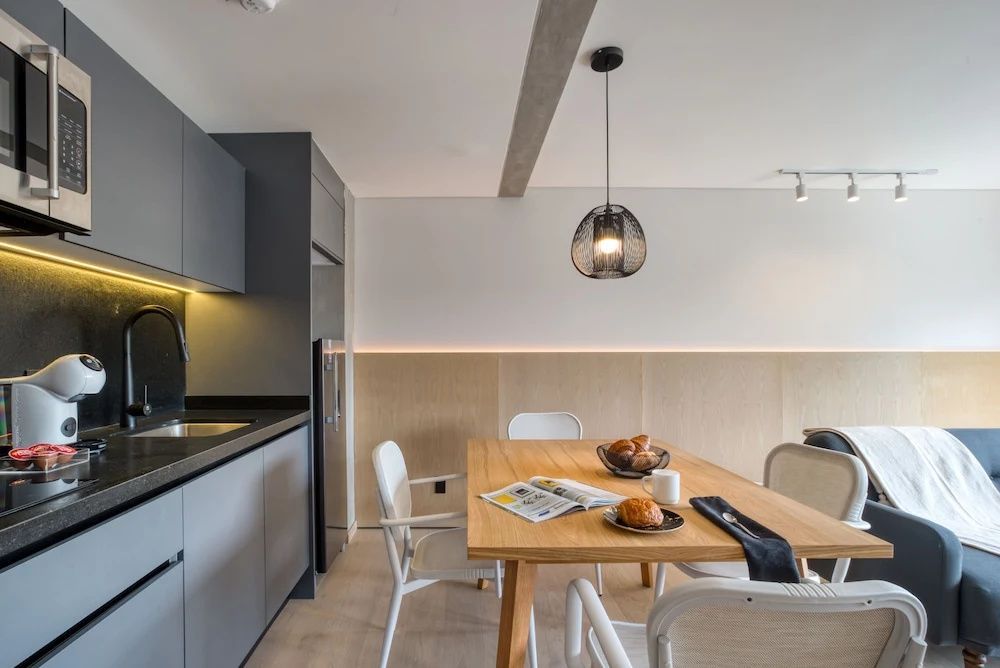 Esstisch aus Holz mit weißen Stühlen neben einer grauen Küchenzeile im hellen Apartment.