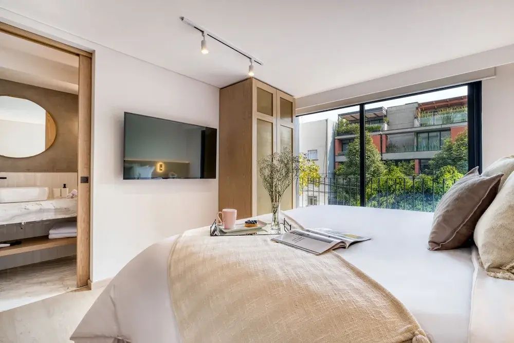 Helles Schlafzimmer mit Wand-TV, Bett und Fenster mit Blick auf begrünte Außenansicht.