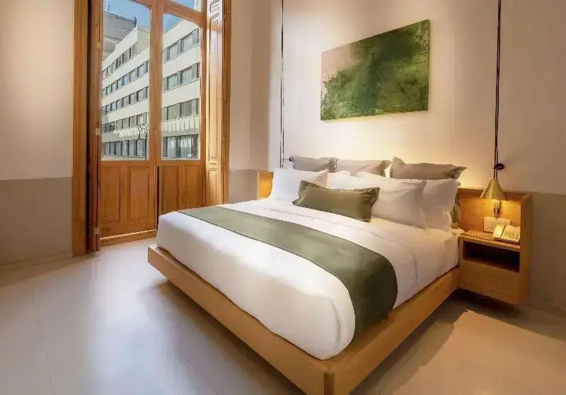 Helles Hotelzimmer mit großem Doppelbett, grünem Bild an der Wand und hohen Fenstern.