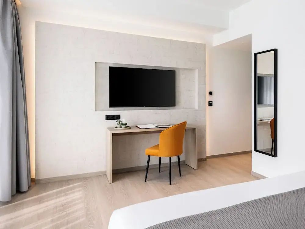 Hell eingerichteter Arbeitsbereich im Hotelzimmer mit Holzschreibtisch, gelbem Stuhl und Fernseher an der Wand.