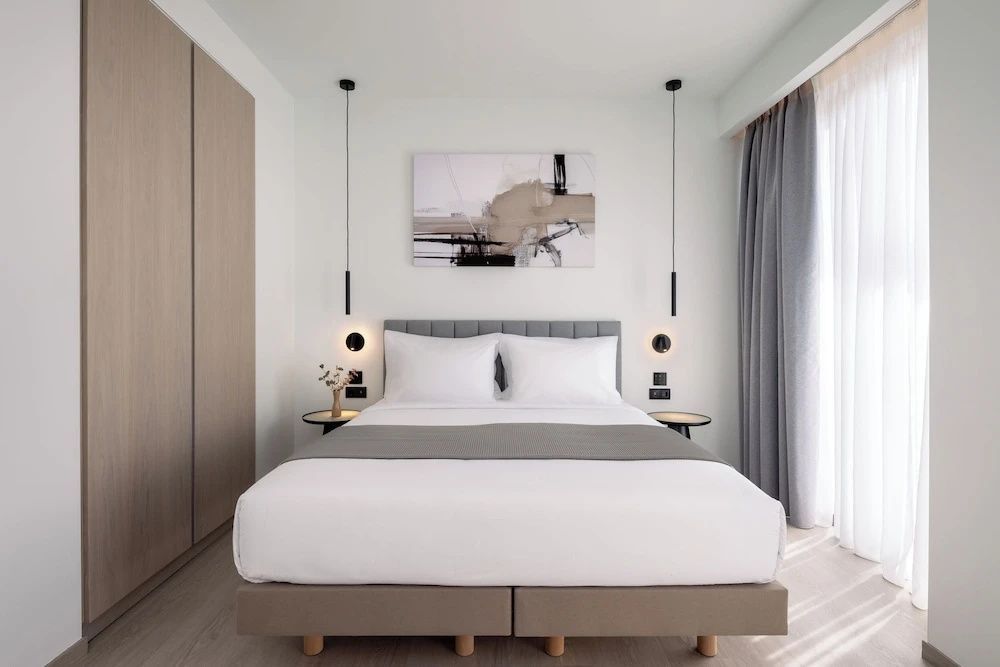 Doppelbett mit weißer Bettwäsche, grauer Tagesdecke und modernen Hängelampen in hellem Hotelzimmer.