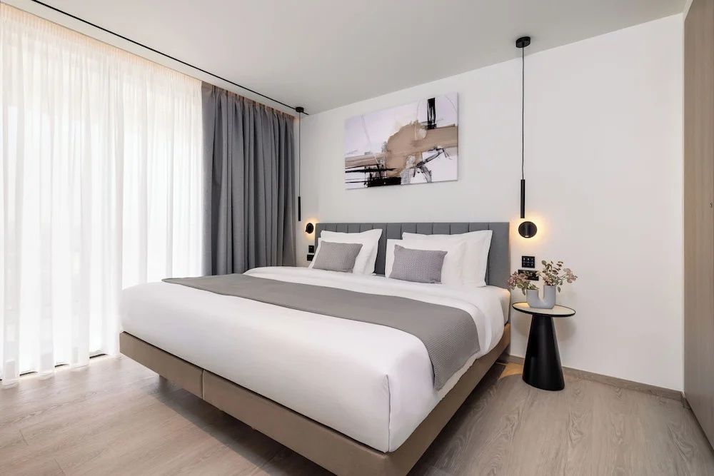 Doppelbett mit grauer Tagesdecke, Stoffkopfteil, Nachttisch und Kunstwerk im Schlafzimmer.