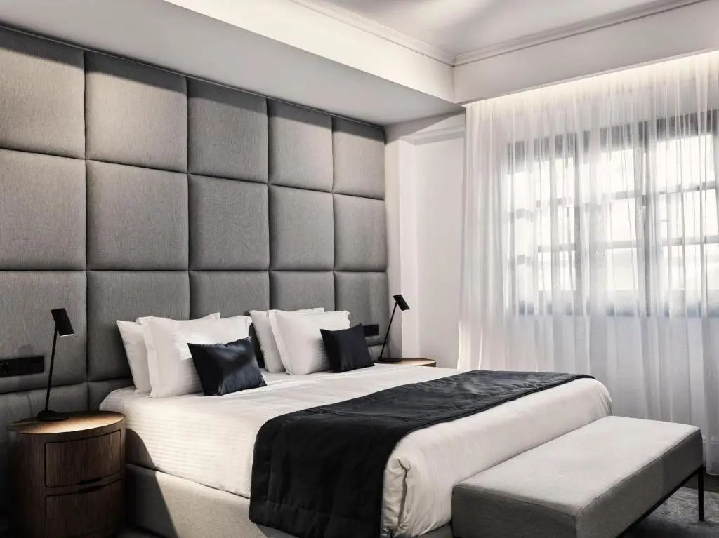 Doppelbett mit grauer Wandpolsterung, weißer Bettwäsche und Vorhang am Fenster.
