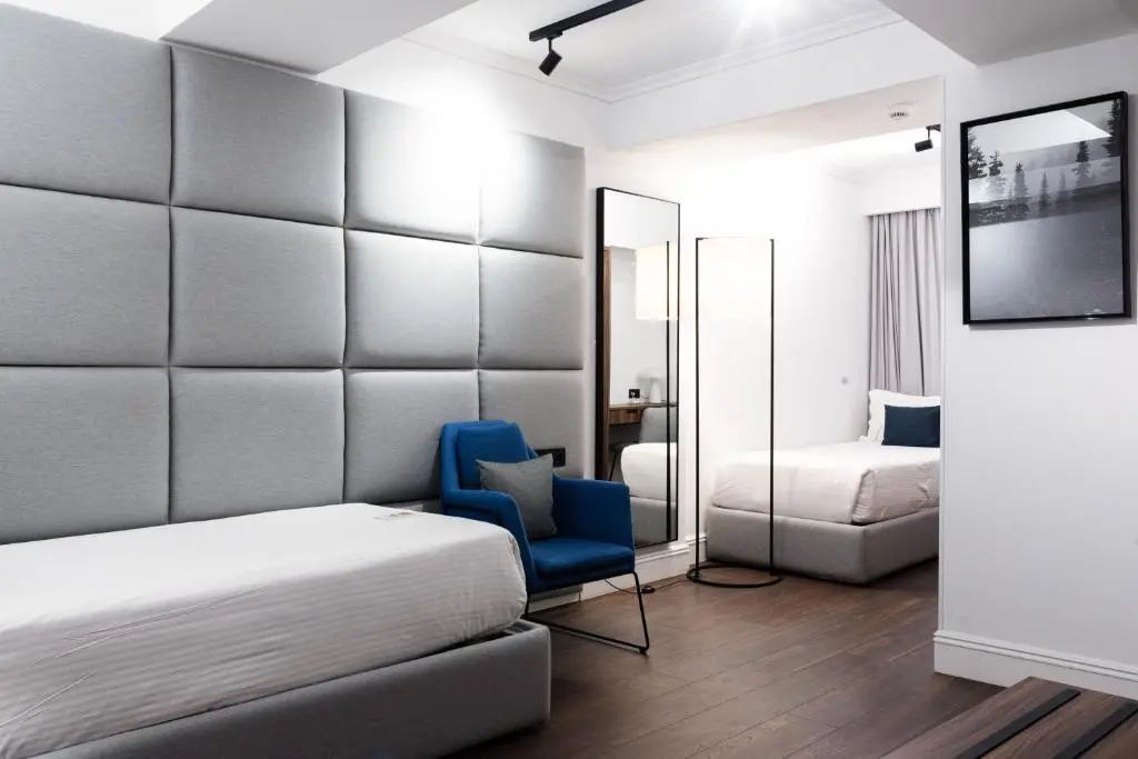 Hotelzimmer mit zwei Einzelbetten, blauem Sessel und stehendem Ganzkörperspiegel.