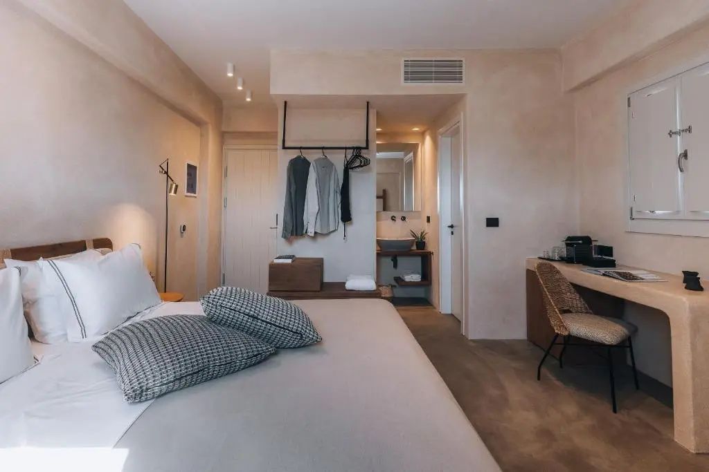 Innenansicht des Hotelzimmers mit Bett im Vordergrund, offener Garderobe und Waschtisch im Hintergrund.