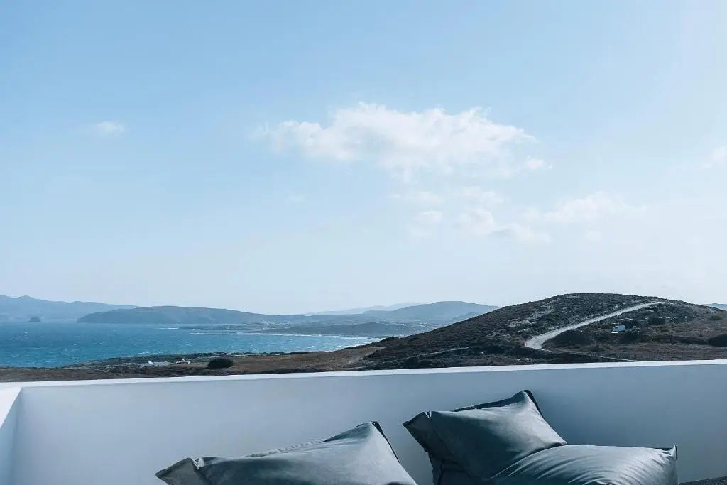 Blick von einer Terrasse mit Sitzkissen auf das Meer und die hügelige Küstenlandschaft unter blauem Himmel.
