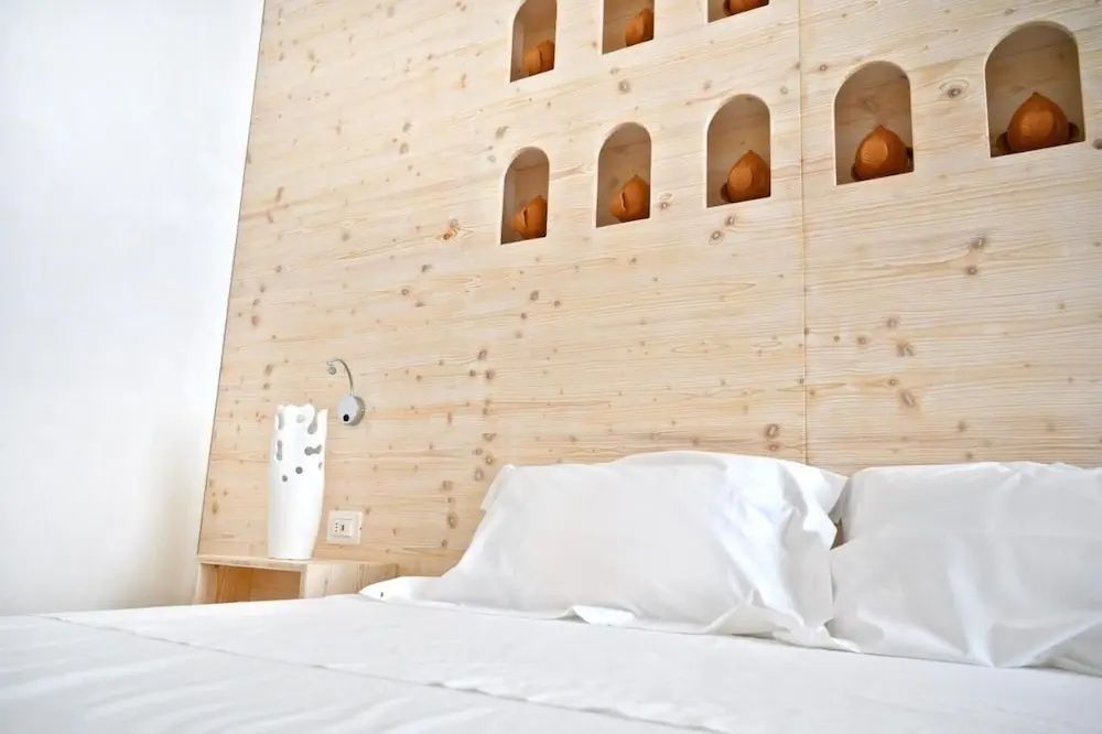 Ein weißes Kissen auf dem Bett und ein Nachttisch mit einer Deko-Vase vor einer Holzwand.