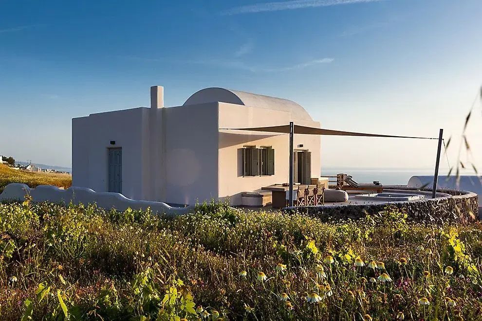 Weißes Ferienhaus mit Tonnendach hinter blühenden Sträuchern unter blauem Himmel.