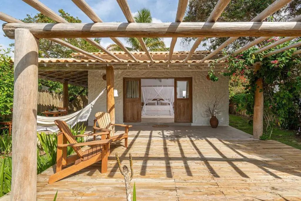 Sonnige Holzterrasse mit Pergola und Stühlen vor einem Bungalow mit offenem Eingang.