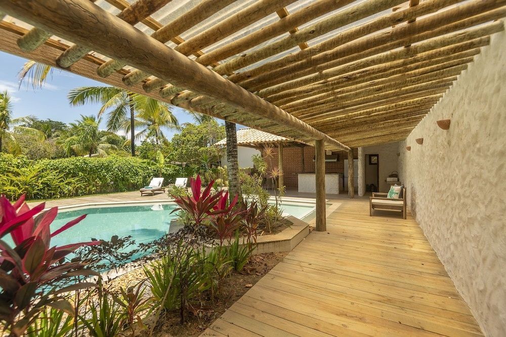 Überdachte Holzterrasse neben dem Poolbereich mit tropischen Pflanzen und Palmen.