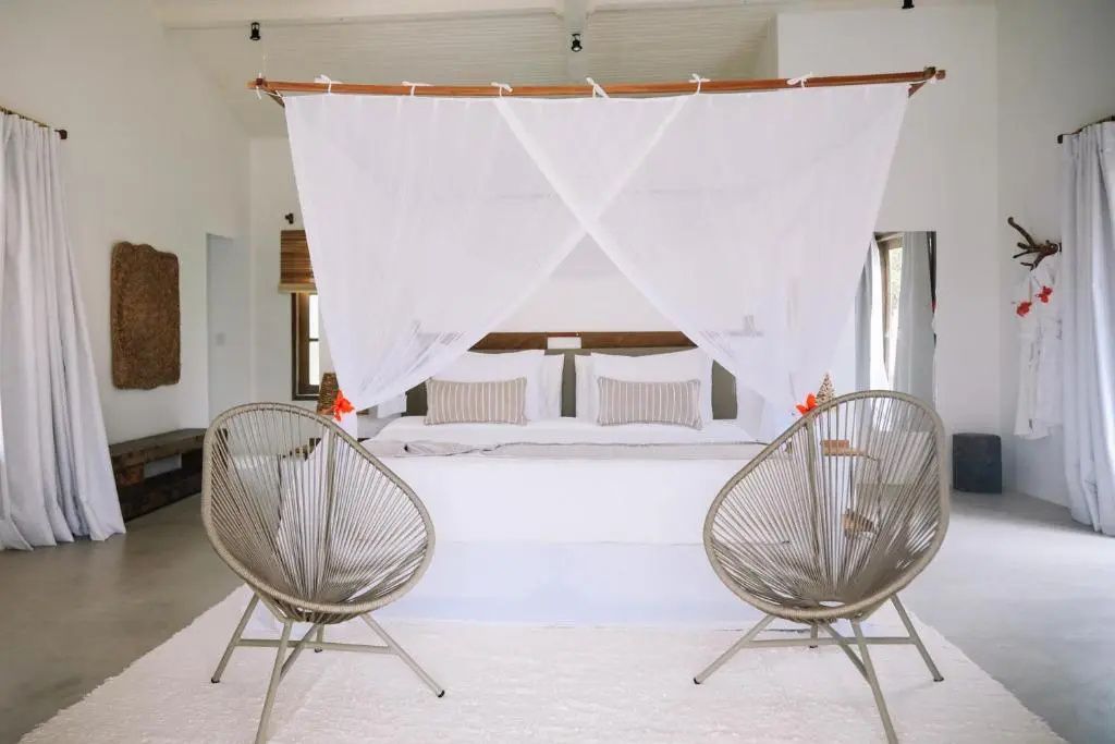 Helles Hotelzimmer mit großem Himmelbett und zwei grauen Sesseln auf weißem Teppich.