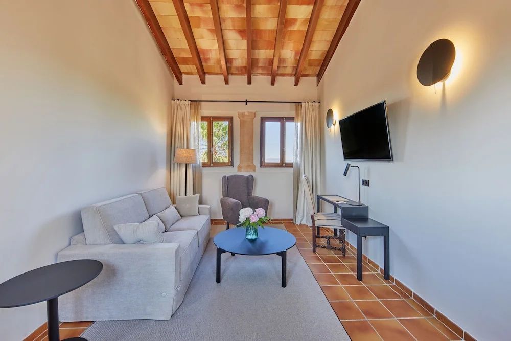 Wohnbereich eines Zimmers mit hellem Sofa, Couchtisch, Sessel und TV unter Holzbalkendecke.