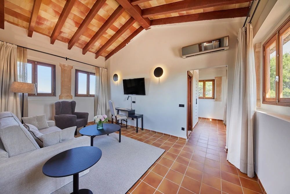 Geräumiges Wohnzimmer im Hotel mit Fliesenboden, Sitzgruppe, Wandfernseher und rustikalen Holzbalken.