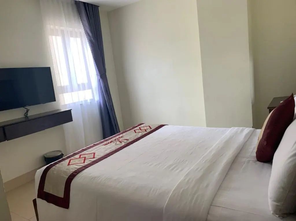 Hotelzimmer mit Doppelbett, Wandfernseher und hellem Fenster mit Gardine.
