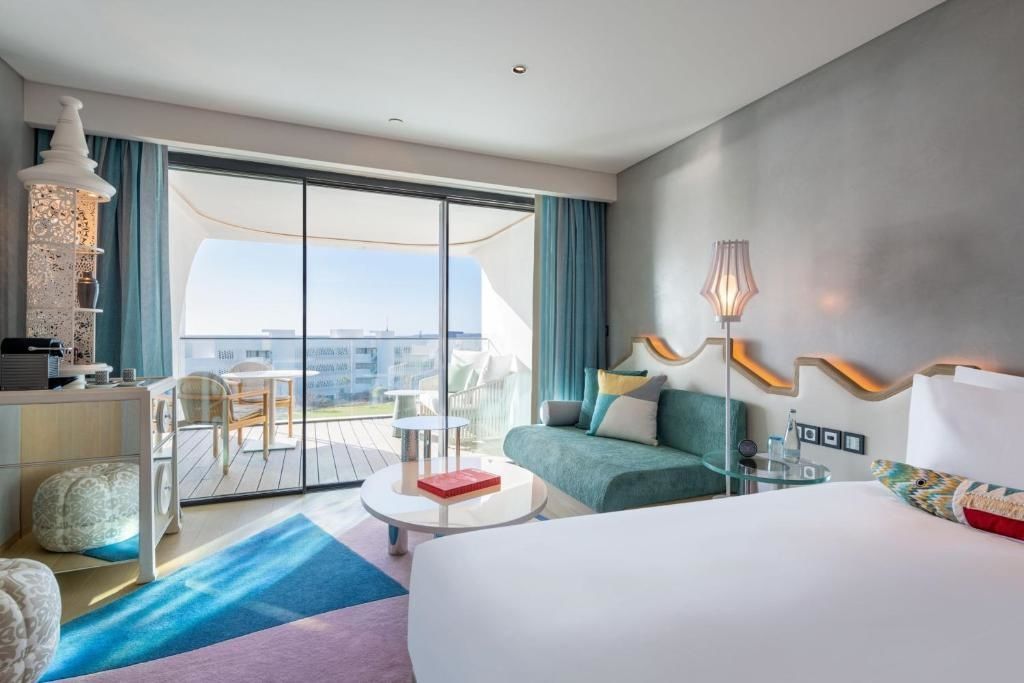 Hotelzimmer mit Bett, grünem Sofa und Schiebetür zum möblierten Balkon.