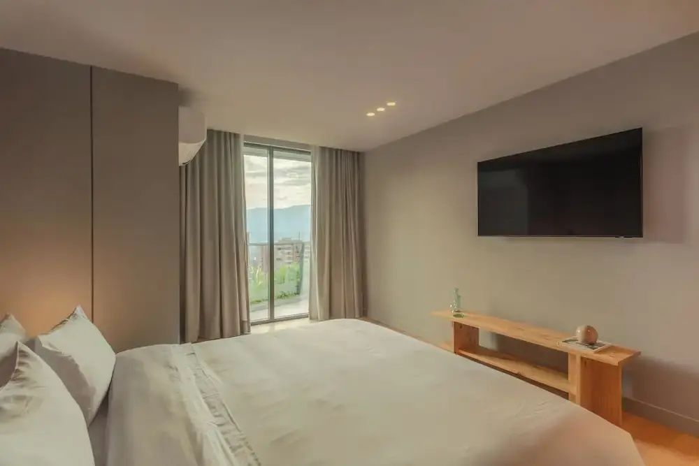 Heller Schlafraum mit weißem Doppelbett, Fernseher an der Wand und Holzregal.