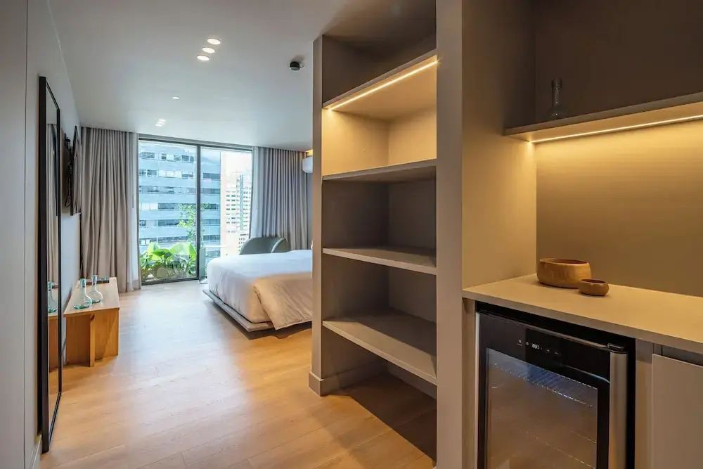Blick vorbei an einem Regal und einer Minibar auf das Bett und die Fensterfront.
