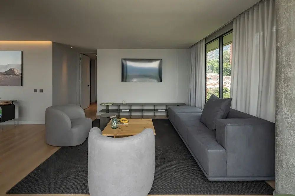 Wohnzimmer mit grauer Couch, zwei Polstersesseln und Wandfernseher.