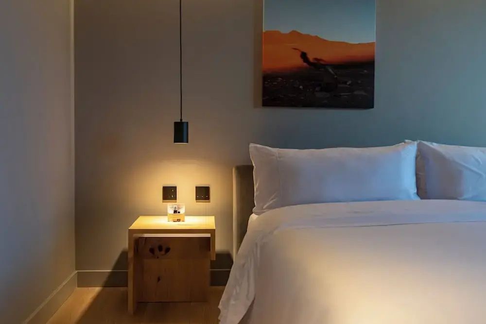 Modernes Nachttischchen aus Holz neben einem weiß bezogenen Doppelbett mit warmer Beleuchtung.