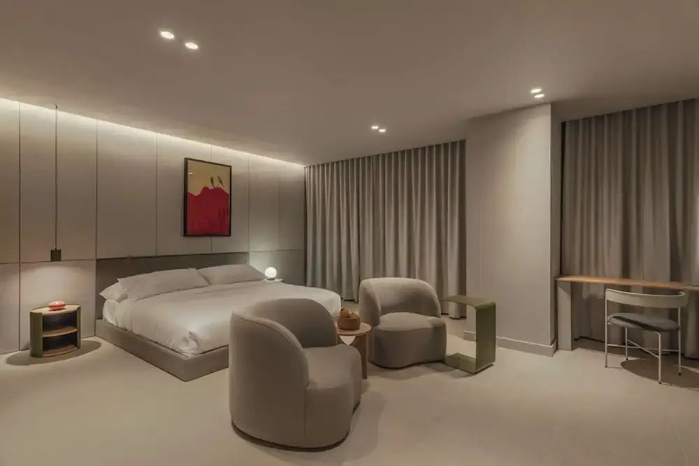 Schlafbereich mit weiß bezogenem Doppelbett, zwei Sesseln und Wandbild in neutralen Tönen.
