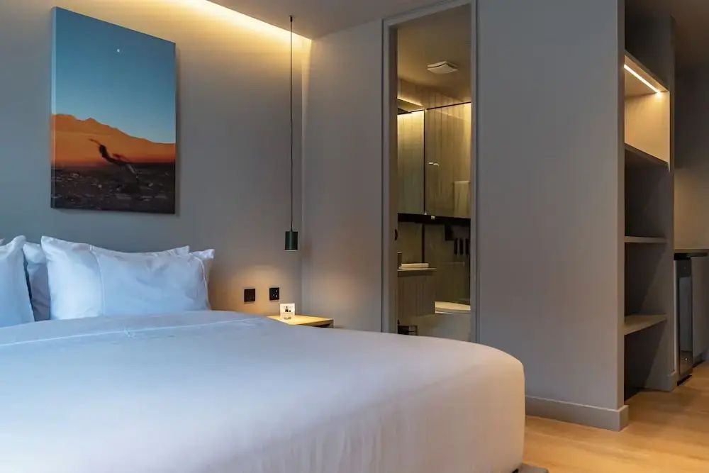 Weißes Doppelbett im Hotelzimmer mit Blick durch eine Glastür in das angrenzende Badezimmer.