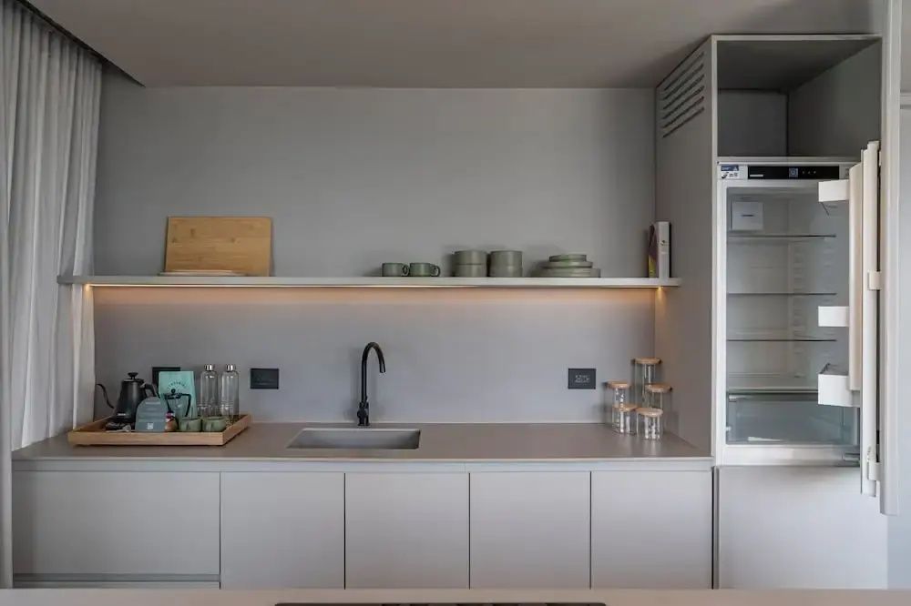 Miniküche mit grauer Arbeitsplatte, Spülbecken, Wandregal mit Geschirr und Kühlschrank.