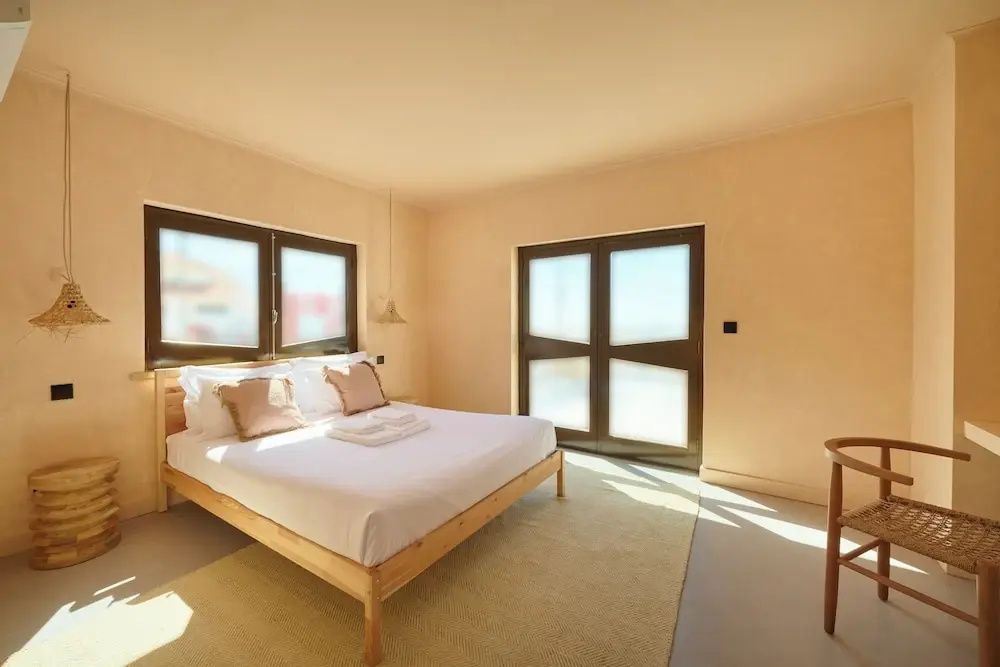 Helles Schlafzimmer mit Holzdoppelbett, beige Wänden und großen Fenstern mit dunklen Rahmen.