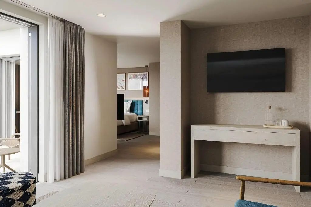 Blick in den Wohnbereich des Zimmers mit hellen Vorhängen, Wandfernseher und Durchgang zum Schlafzimmer.
