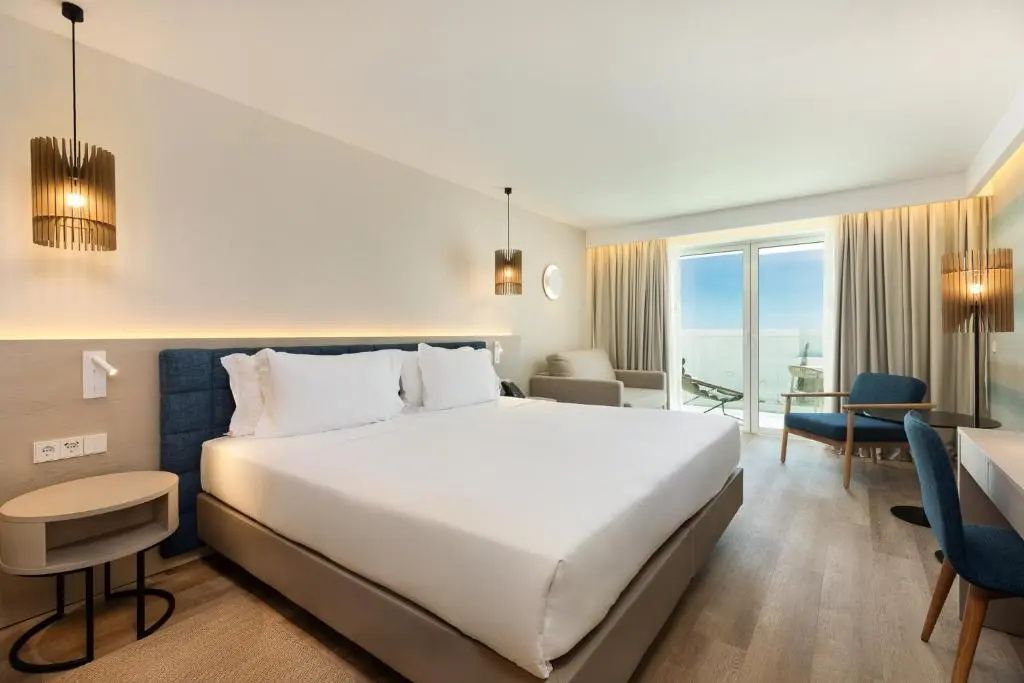 Hell eingerichtetes Hotelzimmer mit Doppelbett, Sitzbereich und Balkonzugang.