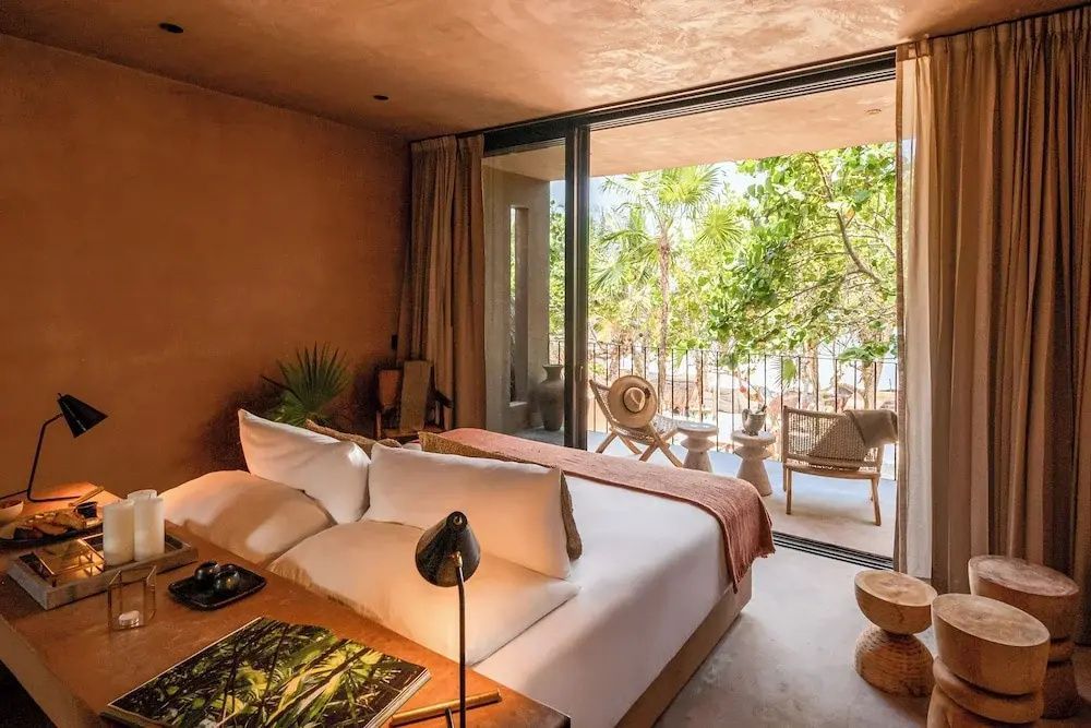 Blick vom Doppelbett auf die private Terrasse mit Stühlen und grünen Bäumen im Hintergrund.