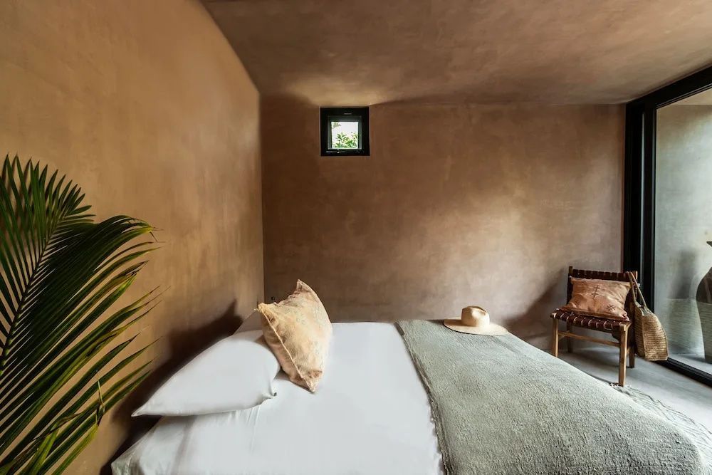 Bett mit weißer Bettwäsche, grüner Decke und Strohhut vor beigefarbenen Lehmwänden mit kleinem Fenster.