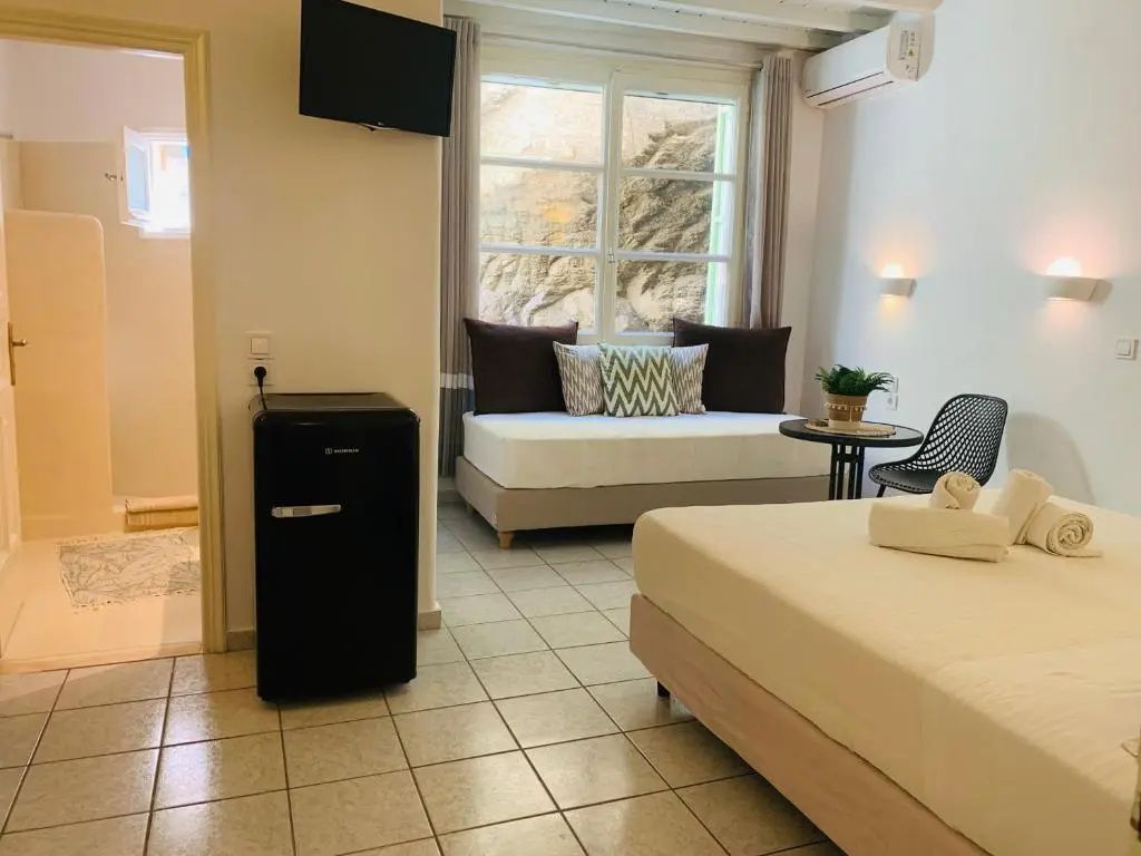 Helles Hotelzimmer mit Bett, Sofa vor dem Fenster, Tisch und kleinem Kühlschrank.