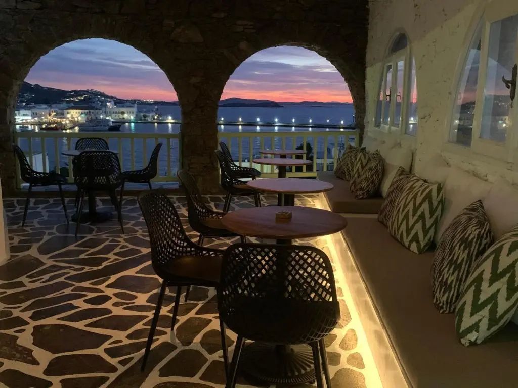 Überdachte Terrasse mit Rundbögen, Tischgruppen und Weitblick auf das Meer bei Abendrot.
