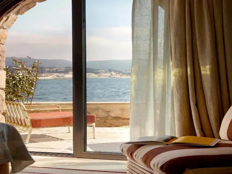 Blick durch eine große Glasschiebetür auf eine Terrasse und das dahinterliegende Meer.
