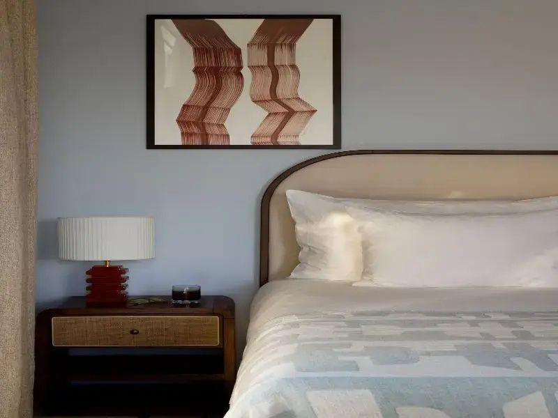 Doppelbett mit weißer Bettwäsche, Holz Nachttisch mit roter Lampe und Kunstwerk an der Wand.
